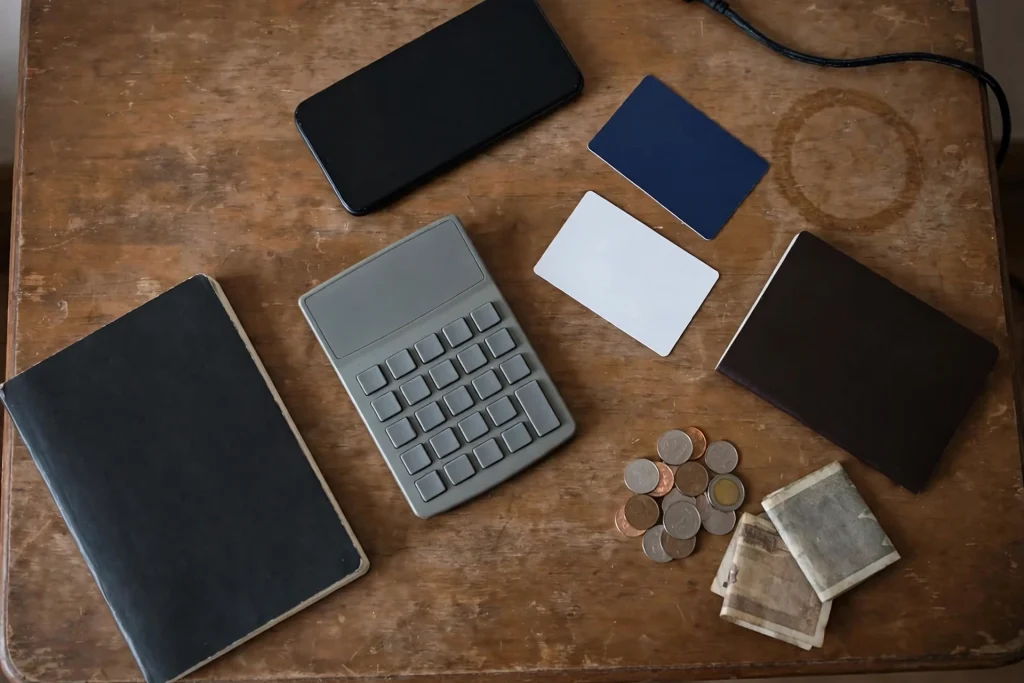 Overhead view of a worn wooden desk with a calculator, closed notebook, face-down phone, two blank payment cards, a passport, coins and banknotes