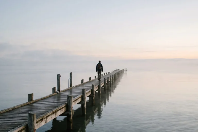 Person walking along a long wooden pier into calm misty sea at dawn