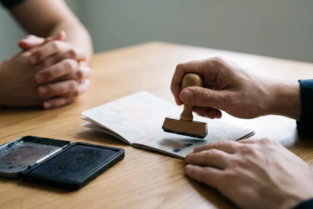 Official hand stamping an open passport page on a wooden desk while an applicant hands wait across the table